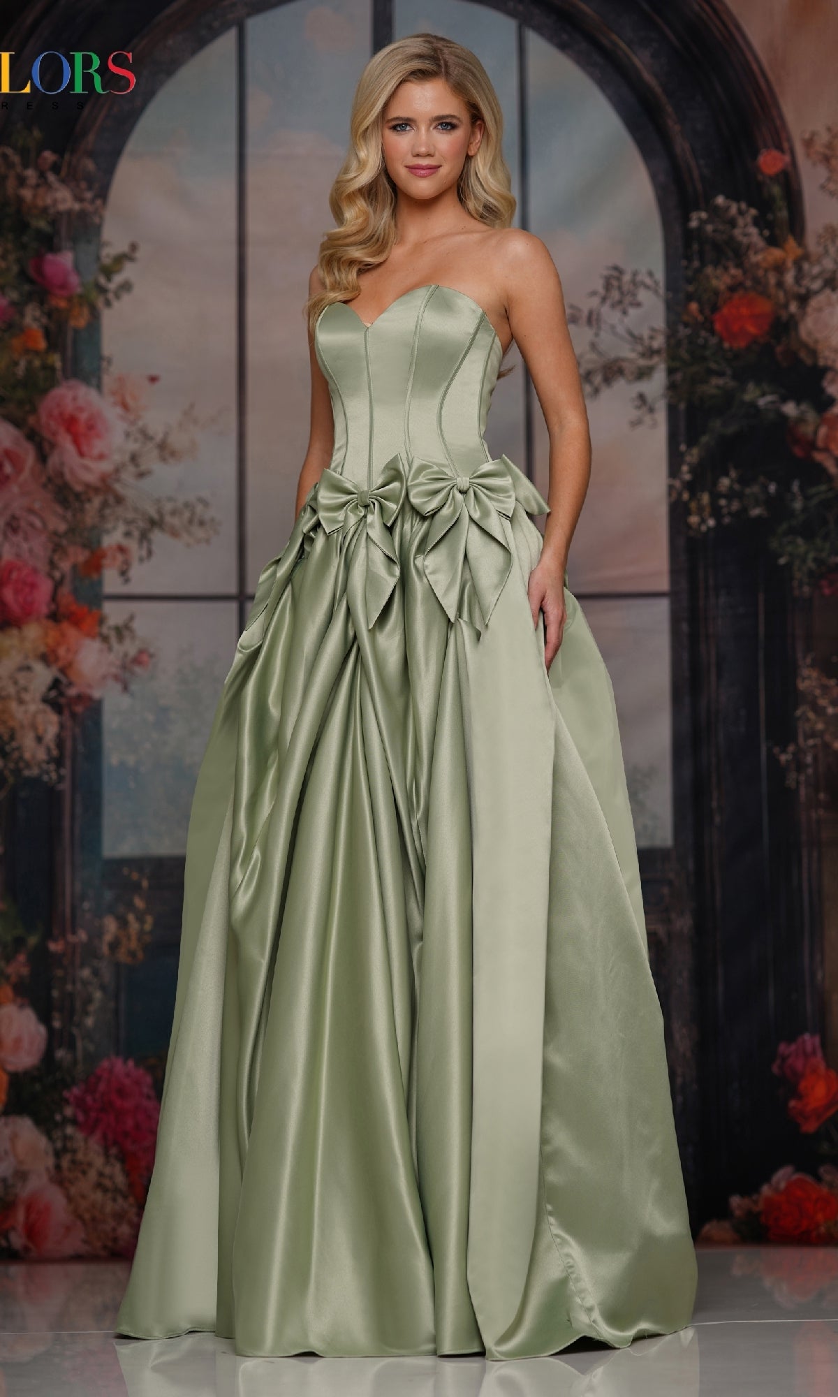 A woman poses by a floral and window backdrop, wearing the Bow-Front Long Prom Ball Gown (Colors Dress 3739) in sage—a strapless sweetheart gown featuring decorative bows and pleated skirt details.